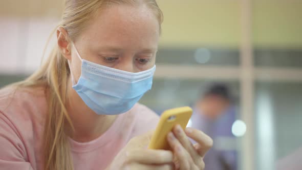 A Young Woman Wearing a Medical Face Mask Sits on a Chair in an Airport Using a Cellphone alt