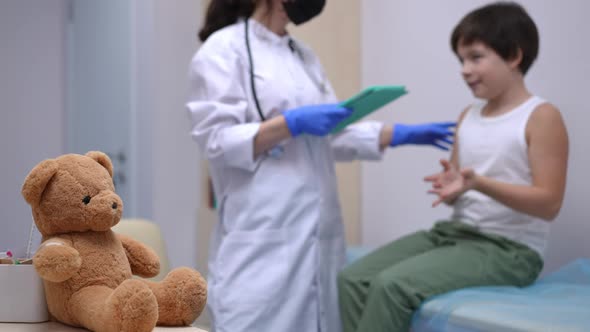 Closeup Brown Teddy Bear in Pediatric Clinic with Blurred Caucasian Boy Talking to Unrecognizable alt