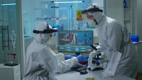 Chemist in Ppe Suit Working at Pc While Lab Technician Bringing Her Blood Samples alt