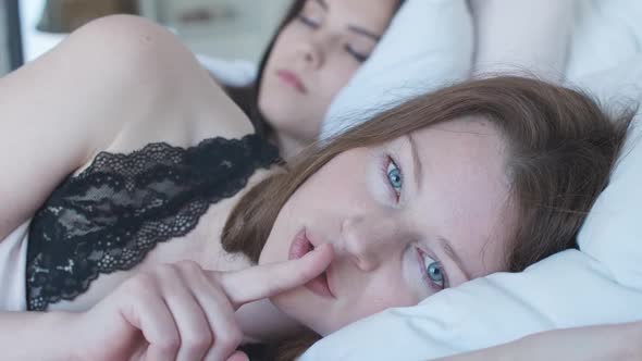 Close-up Portrait of Blue-eyed Redhead Woman Showing Hush Gesture As Lying in Bed with Brunette alt