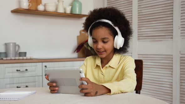 Little African American Girl in Big White Headphones Plays a Game Using a Digital Tablet alt