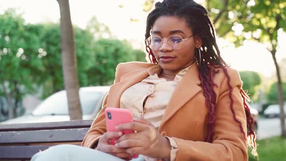 Young African American Woman Sitting on a Bench in the City and Using Her Smartphone alt
