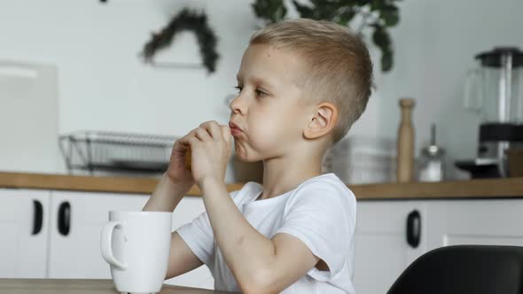 A Blonde Boy Child Eats Lunch in A Bright Kitchen, Eats A Pie And Drinks Tea or Coffee or Cocoa alt