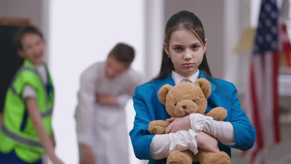 Sad Stressed Bullied Girl Hugging Teddy Bear As Boys Laughing Pointing at Background alt