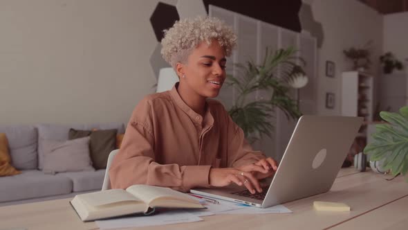 Smiling Young African Woman Looking at Laptop and Book Learning at Home alt
