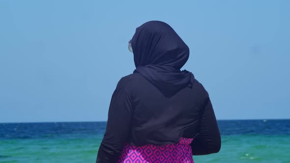 a Woman in a Hijab is Standing with Her Back to the Camera Against the Background of the Sea and alt