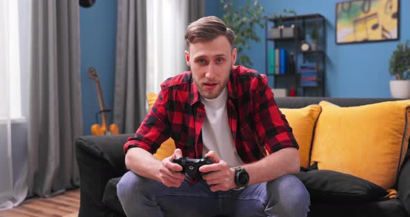 Cheerful American Man Resting in Room Sitting on Sofa Playing Video Games Using Joysticks alt
