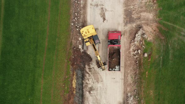 Top View Yellow Excavator Picks Up Land From the Field and Loads It Onto a Red Truck alt