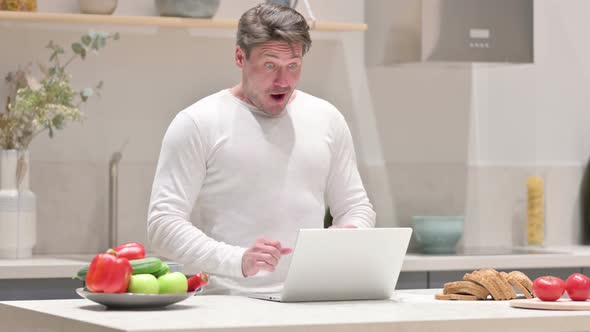 Middle Aged Man Celebrating Success While Using Laptop in Kitchen alt