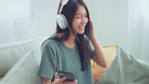 Happy young woman wear wireless headphones having fun listen music in smartphone and sit on sofa alt