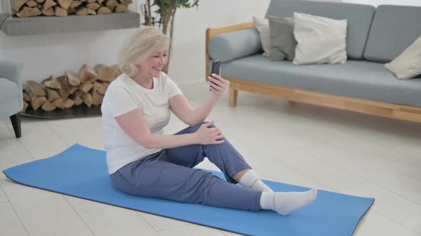 Old Woman Talking on Video Call on Smartphone While Sitting on Yoga Mat alt