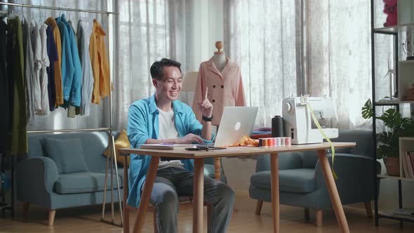 Male Designer Working On A Laptop And Thinking Then Raising Index Finger While Designing Clothes alt