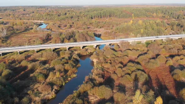 The car drives over the bridge over the river next to the autumn forest. Aerial view