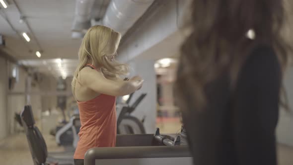 Positive Athletic Blond Woman Walking on Treadmill and Talking with Unrecognizable Female Caucasian alt