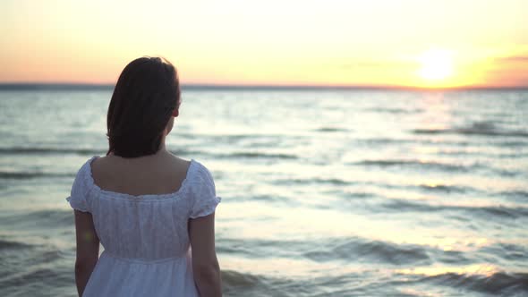 Attractive Young Woman Looks at the Sunset on the Beach By the Sea. The Girl in a White Dress Stands alt