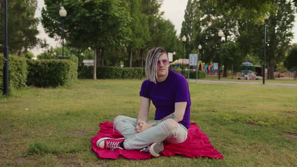 A handsome guy with dreadlocks in casual clothes, sitting on a plaid in a park on a green lawn alt