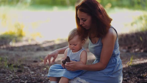 mother and baby sit in a park or in a forest on the ground looking at twigs and cones alt