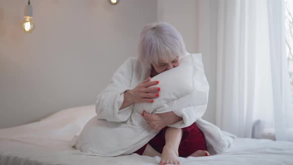Happy Smiling Mature Woman Smelling Fresh Clean Pillow Looking at Camera Sitting on Bed alt