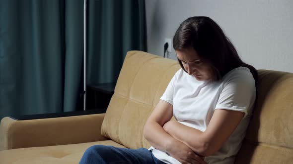 Young Woman Hunched Over Holding Belly While Sitting on the Couch alt