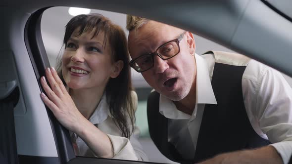 Shooting From Car Salon of Positive Adult Successful Caucasian Couple Talking Choosing New Vehicle alt