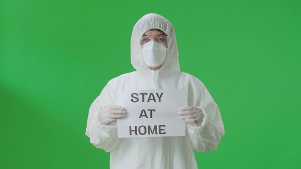 Asian Man Wear Protective Uniform PPE And Holding Stay At Home Sign In The Green Screen Studio alt