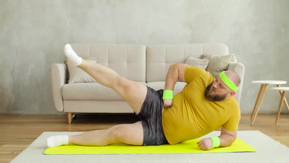 Fat Man in Yellow Sportswear Doing Legs Lifting Exercises Lying on Mat at Home