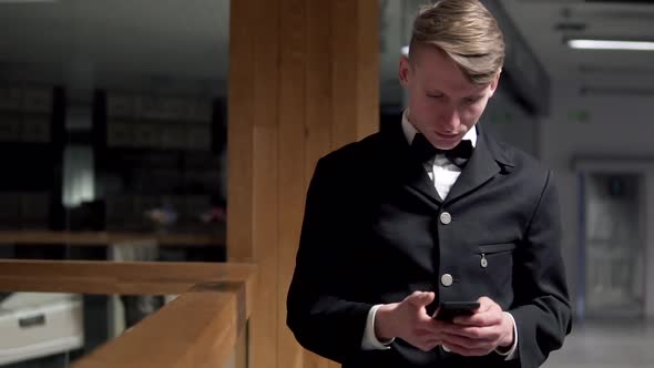 Young Attractive Businessman Standing Near the Stairs and Uses a Smart Phone alt
