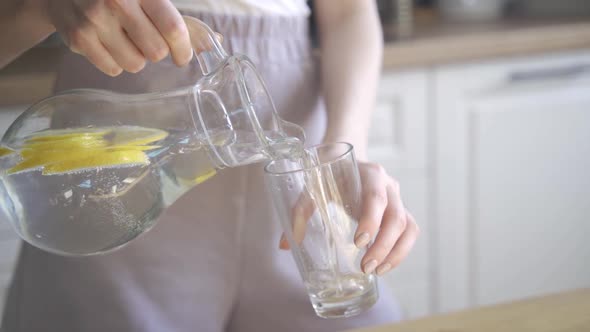 A Young Blonde Woman Pours Refreshing Water with Lemon Into a Transparent Glass and Drinks It alt