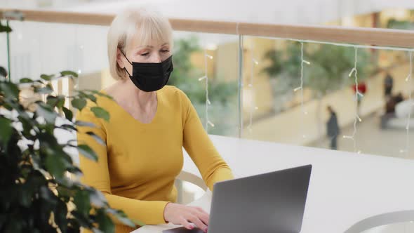 Mature Woman Professional Worker Freelancer in Protective Mask Sitting in Workplace Typing on Laptop alt