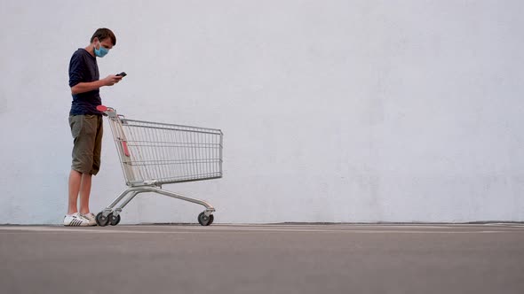 Man in Mask with Smartphone Pushing Shopping Cart. Customer with Shopping Trolley Along the Wall of alt