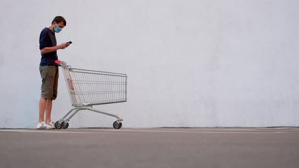 Man in Mask with Smartphone Pushing Shopping Cart. Customer with Shopping Trolley Along the Wall of