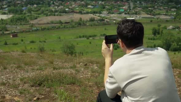 A Young Adult Male Takes a Smartphone Picture From the Top of a Large Hill alt