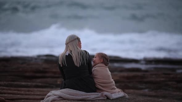 Back View of Blond Woman with Little Girl Cuddling in Plaid on Rocky Shoreline in Twilight Time