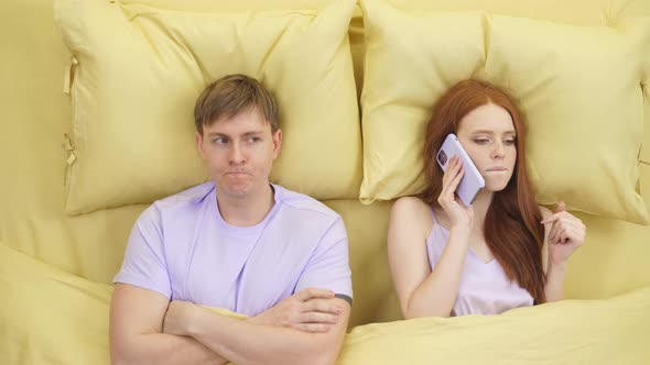 Young Beautiful Couple in Bed Woman is Busy Talking on Phone  Man Upset