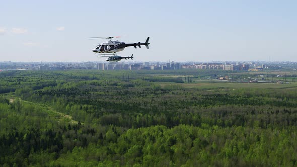 Two Modern Bell Helicopters with White and Black Coloring Fly in Parallel Over Highway Green Trees