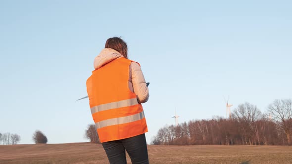 Female Engineer in Orange Vesta Goes to Wind Turbines with a Tablet to Checks Their Operation alt