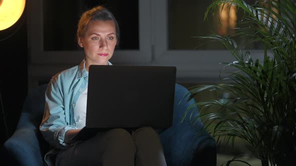 Woman is Sitting in the Armchair and Working on a Laptop at Night alt