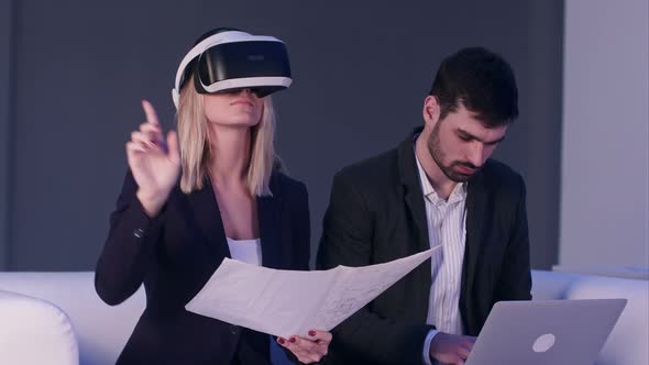 Blonde Woman in VR Headset Holding Construction Plans While Her Male Partner Typing on the Laptop alt