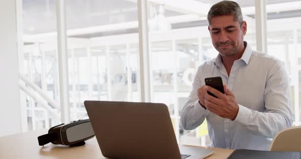 Businessman using mobile phone while working on laptop in modern office
