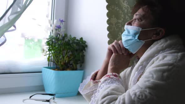 Old Despair Woman in Protective Medical Mask Looking Sadly Out Window Sitting at Home in Quarantine
