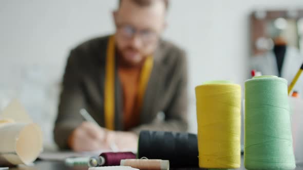 Fashion Desigher Drawing in Tailoring Studio at Work Table with Sewing Materials alt
