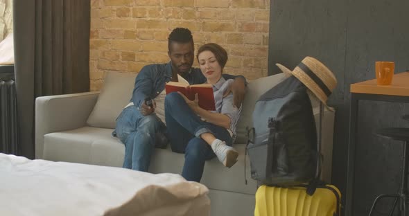 Wide Shot of Relaxed Interracial Couple Sitting on Couch with Remote Control and Book. Packed alt