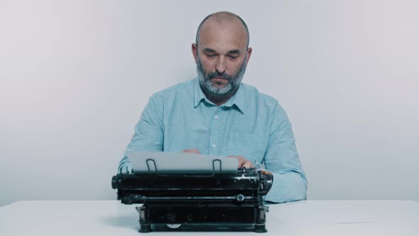 Male Writer Works Sitting at the Table. A Man Writes on Paper, Typewriter Is in Front of Him alt
