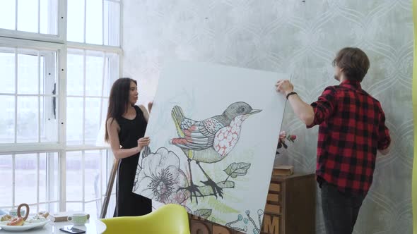 Happy couple hanging picture on the wall at home, A Young Couple In Their Bright Apartment  alt