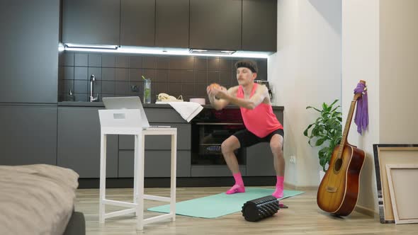 Funny man athlete in pink top doing squats watching online fitness classes on notebook at home alt
