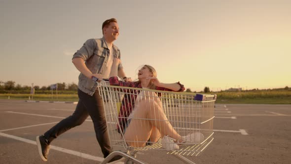 Young Stylish Coupe Having Fun Riding with Shopping Cart on the Outdoor Parking Near the Supermarket alt
