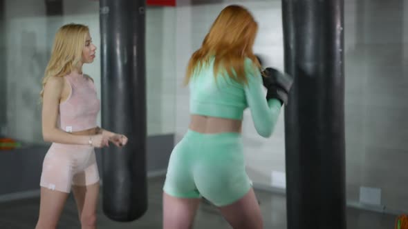 Back View of Redhead Motivated Strong Woman Boxing Punching Bag As Blond Coach Talking Endorsing alt