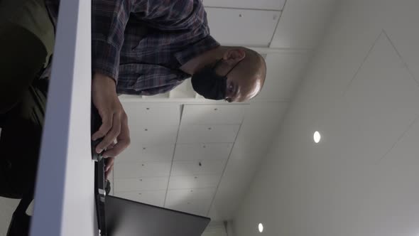 Adult Male On Laptop Wearing Face Mask On Large Desk. Vertical Video, Locked Off, Low Angle alt