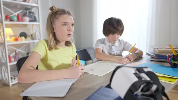 Portrait of Focused Genius Girl Thinking and Writing in Workbook with Blurred Boy Drawing alt