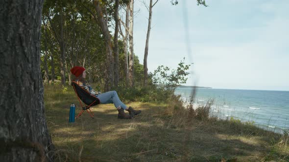 Female traveler sitting on camping chair and admires ocean view
